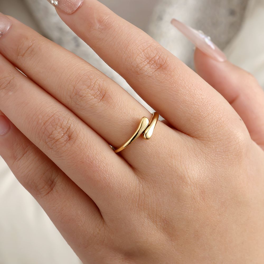 Ein goldfarbener, filigraner Ring in offener Tropfenform wird an einem Finger getragen. Die Enden des Rings wirken wie zwei fließende Tropfen, die sich fas berühren.