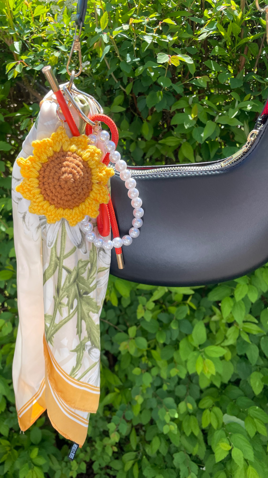 Ein sommerliches Accessoire-Set, bestehend aus einem cremefarbenen Seidentuch mit gelber Kante und Sonnenblumenmuster, einer gehäkelten Sonnenblume, einem roten Band in Schleifenform und einer Perlenkette, ist dekorativ an einer schwarzen Handtasche befestigt. Die Accessoires hängen an einem goldfarbenen Karabiner or einem grünen Busch im Sonnenlicht.