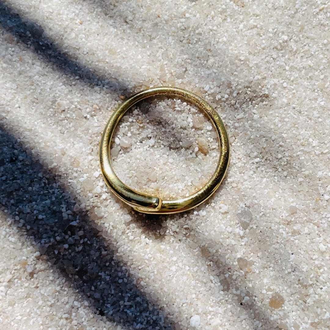 Ein glänzender, goldfarbener Ring mit offener, minimalistischer Tropfenform liegt flach auf hellem Sand. Über den Sand ziehen sich weiche Schatten von Palmenblättern, was eine sommerliche, entspannte Atmosphäre schafft.