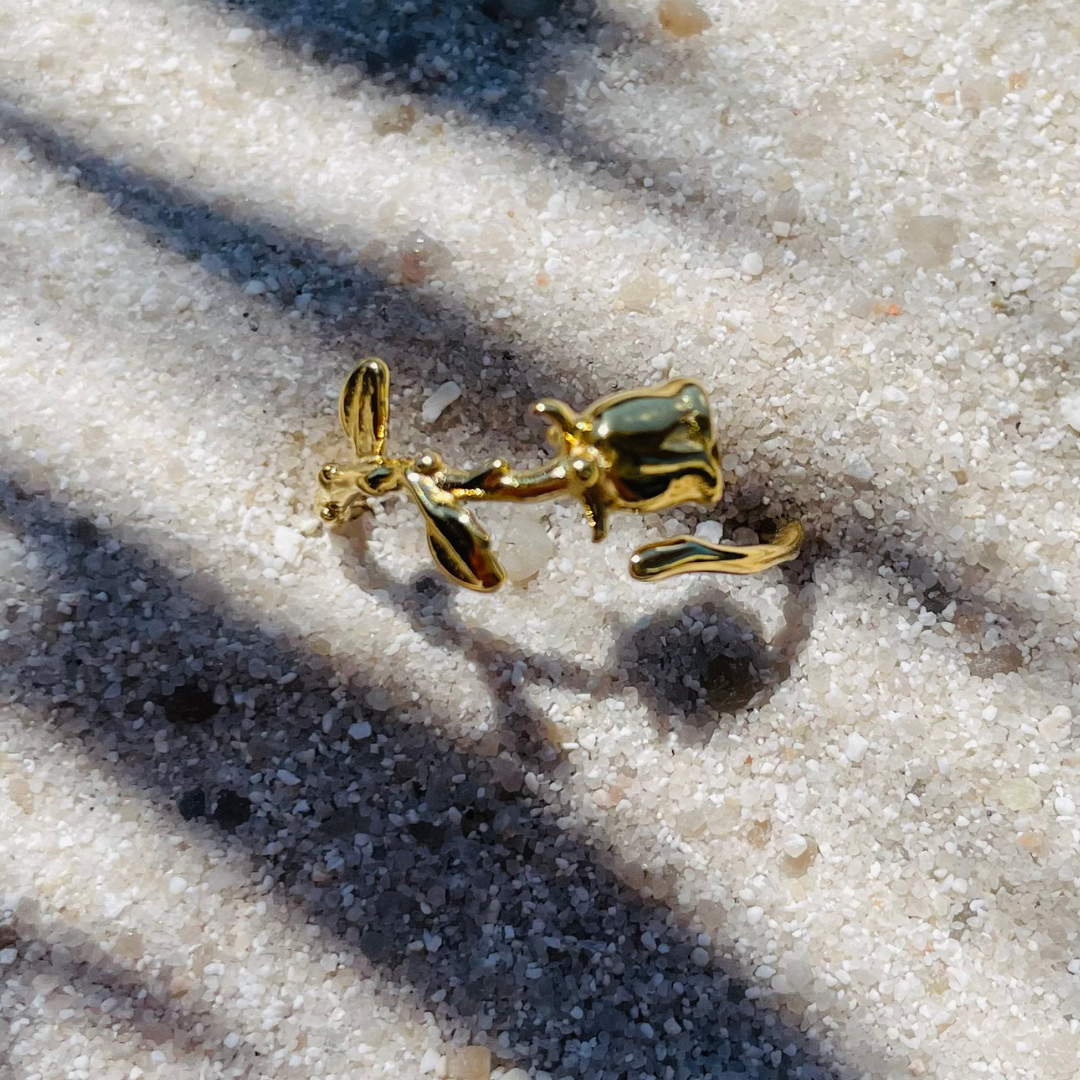 Ein goldfarbener Ring in Form einer Rose liegt auf hellem, feinkörnigem Sand. Die Sonne wirft Schatten von Palmenblättern über das Bild, wodurch eine tropische, entspannte Stimmung entsteht.