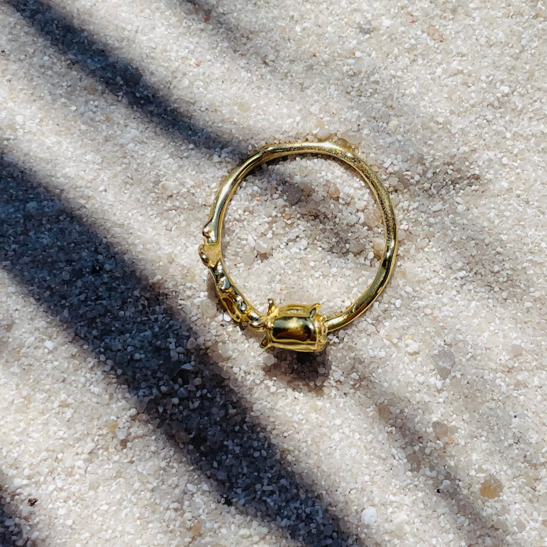 Ein goldfarbener Ring in Form einer Rose liegt flach auf hellem, feinkörnigem Sand. Die Sonne wirft Schatten von Palmenblättern über das Bild, wodurch eine tropische, entspannte Stimmung entsteht.