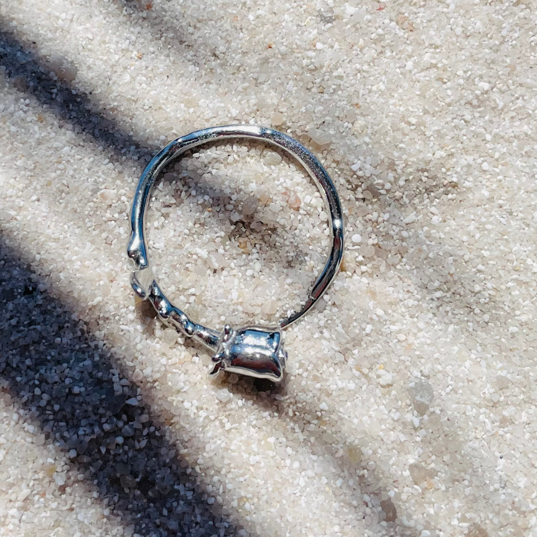 Ein silberfarbener Ring in Form einer Rose liegt flach auf hellem, feinkörnigem Sand. Die Sonne wirft Schatten von Palmenblättern über das Bild, wodurch eine tropische, entspannte Stimmung entsteht.