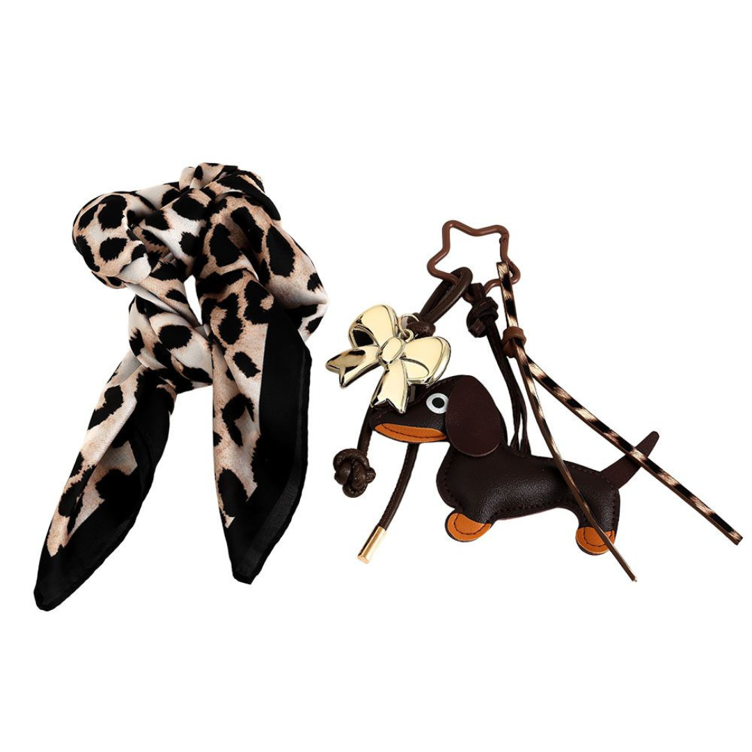 Ein stilvolles Bag Charm Set mit Leopardenmuster-Tuch, Schleifenanhänger in Gold, sternförmigen Karabiner und einem dunkelbraunen Dackelanhänger mit braunen Pfoten. Die Elemente sind farblich aufeinander abgestimmt und ergeben ein verspieltes Accessoire-Set.