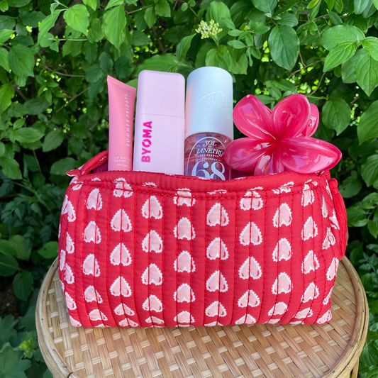 Rote Kosmetiktasche mit rosa Herzmuster, gefüllt mit pinkfarbenen Beautyprodukten und einer glänzenden, rosafarbenen Haarklammer in Blütenform. Aufgenommen auf einem runden Rattantisch vor grünem Blattwerk.