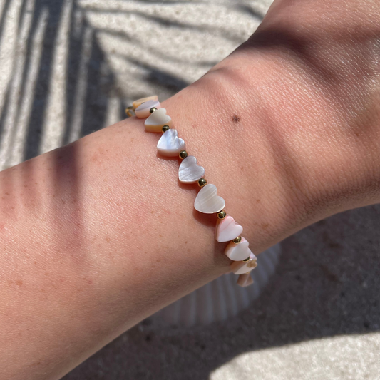 Nahaufnahme eines Handgelenks mit einem handgemachten Armband aus herzförmigen Muschelperlen in zarten Rosatönen. Zwischen den Muschelperlen sind kleine silberne Kugeln. Die Perlen glänzen leicht in der Sonne, im Hintergrund ist Sand mit Muschelschale. und Schattenwurf zu sehen.