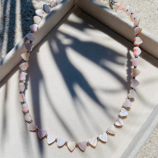 Handgemachte Halskette aus kleinen, herzförmigen Muschelperlen in Rosa mit kleinen silbernen Kugeln dazwischen. Die Kette liegt auf einem hellen Tablett, umgeben von Sand, Sonnenlicht und sanften Schatten.