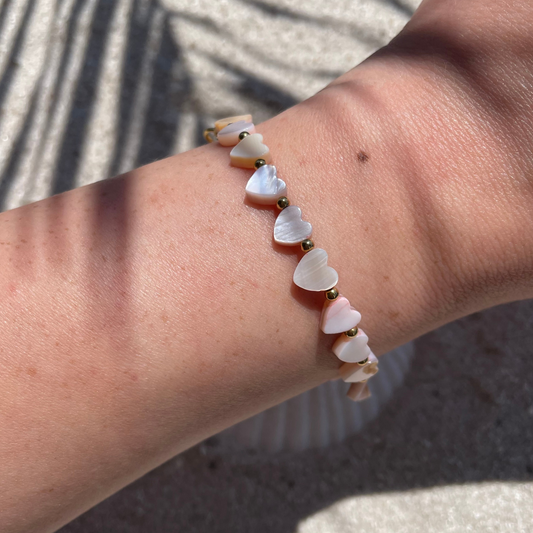 Nahaufnahme eines Handgelenks mit einem handgemachten Armband aus herzförmigen Muschelperlen in zarten Rosatönen, dazwischen sind kleine goldene Kugeln. Die Perlen glänzen leicht in der Sonne, im Hintergrund ist Sand mit Muschelschale und Schattenwurf zu sehen.