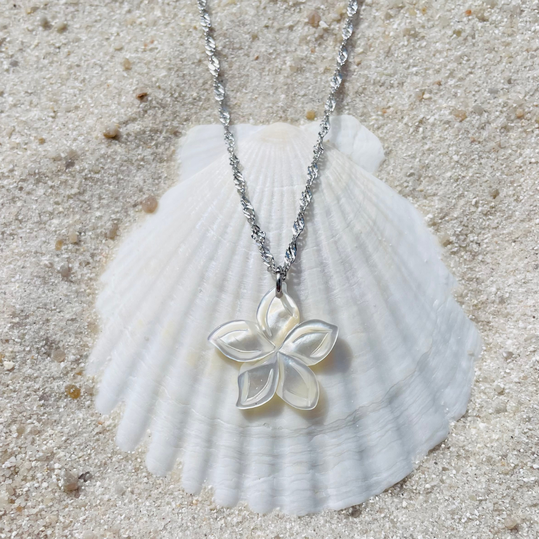 Silberkette mit einem weißen Blütenanhänger aus schimmernder Muschelperle liegt dekorativ auf einer weißen Muschelschale im Sand. 