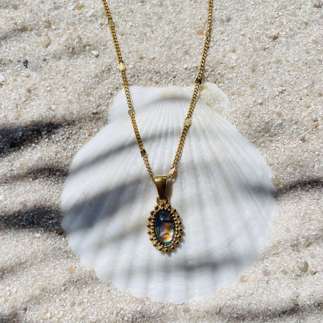 Eine goldfarbene Halskette mit einem ovalen Abalone-Perlmuttanhänger liegt auf einer weißen Muschel im Sand. Der Anhänger ist mit feinen goldenen Details eingefasst. Sonnenlicht und Palmenschatten erzeugen eine sommerliche Atmosphäre. 