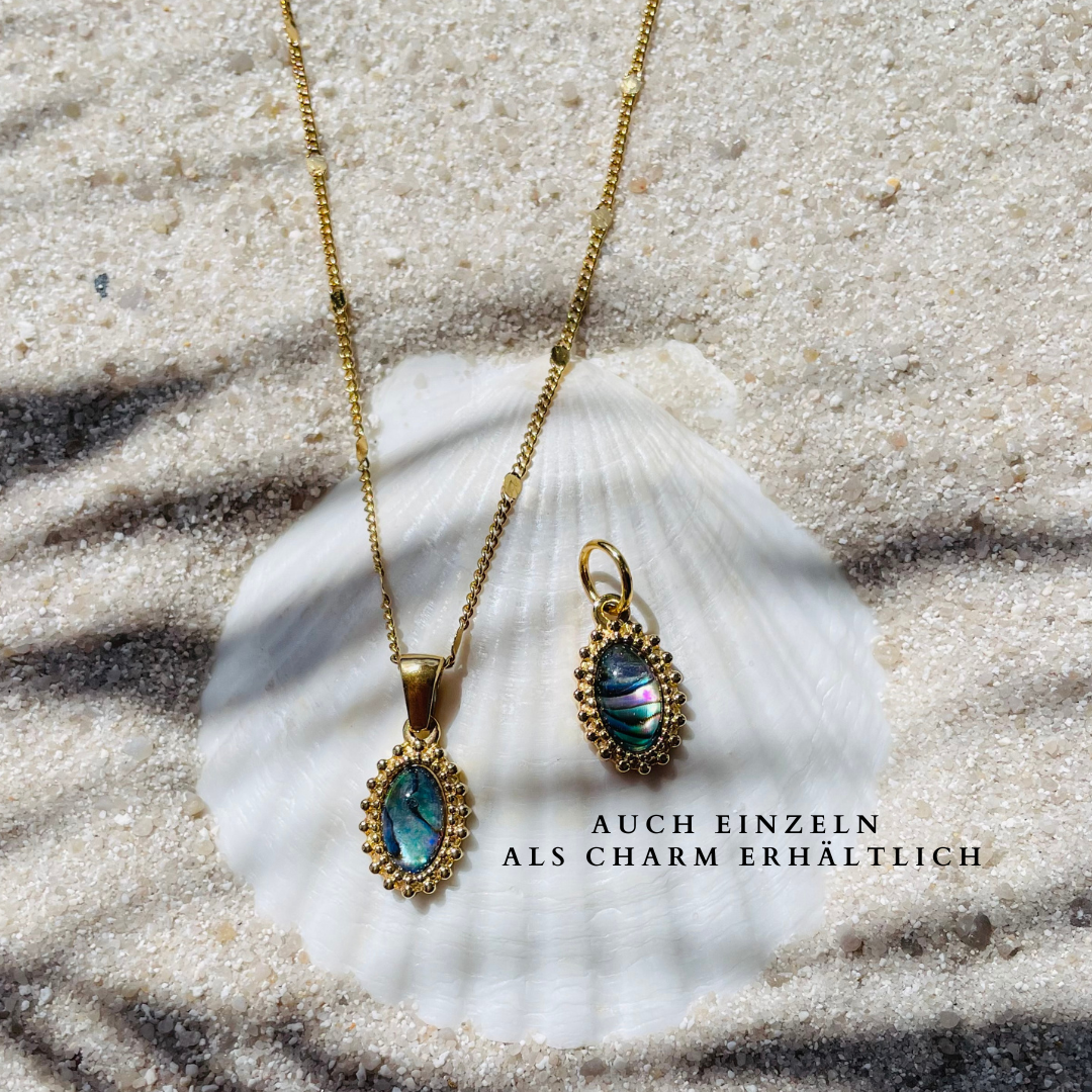 Eine goldfarbene Halskette mit einem ovalen Abalone-Perlmuttanhänger und ein einzelner Charm aus demselben Material,  liegen auf einer weißen Muschel im Sand. Die Anhänger sind mit feinen goldenen Details eingefasst. Sonnenlicht und Palmenschatten erzeugen eine sommerliche Atmosphäre. Unter dem Charm steht als Hinweis der Text: "Auch einzeln als Charm erhältlich. 