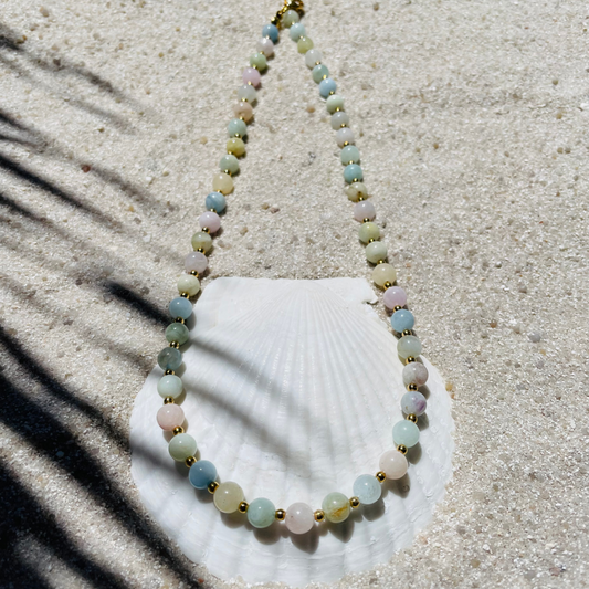 Handgefertigte Halskette aus runden, polierten Beryll-Perlen in zarten Pastellfarben - rosa, grün, blau und gelb - mit kleinen goldenen Kugeln dazwischen. Fotografiert auf hellem Sand mit einer weißen Muschel im Hintergrund und sanften Schattenspielen. 