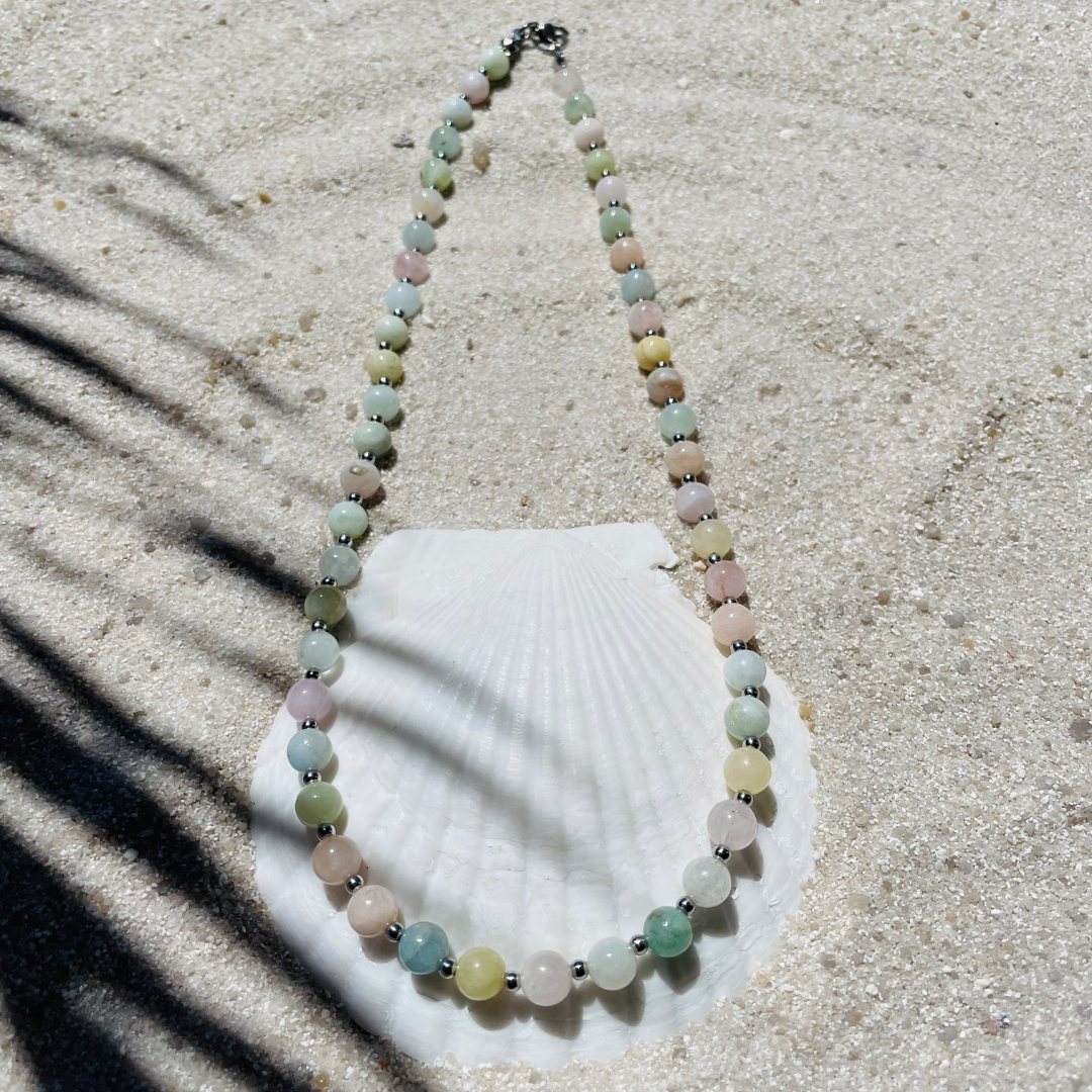 Handgefertigte Halskette aus runden, polierten Beryll-Perlen in zarten Pastellfarben - rosa, grün, blau und gelb - mit kleinen silbernen Kugeln dazwischen. Fotografiert auf hellem Sand mit einer weißen Muschel im Hintergrund und sanften Schattenspielen. 