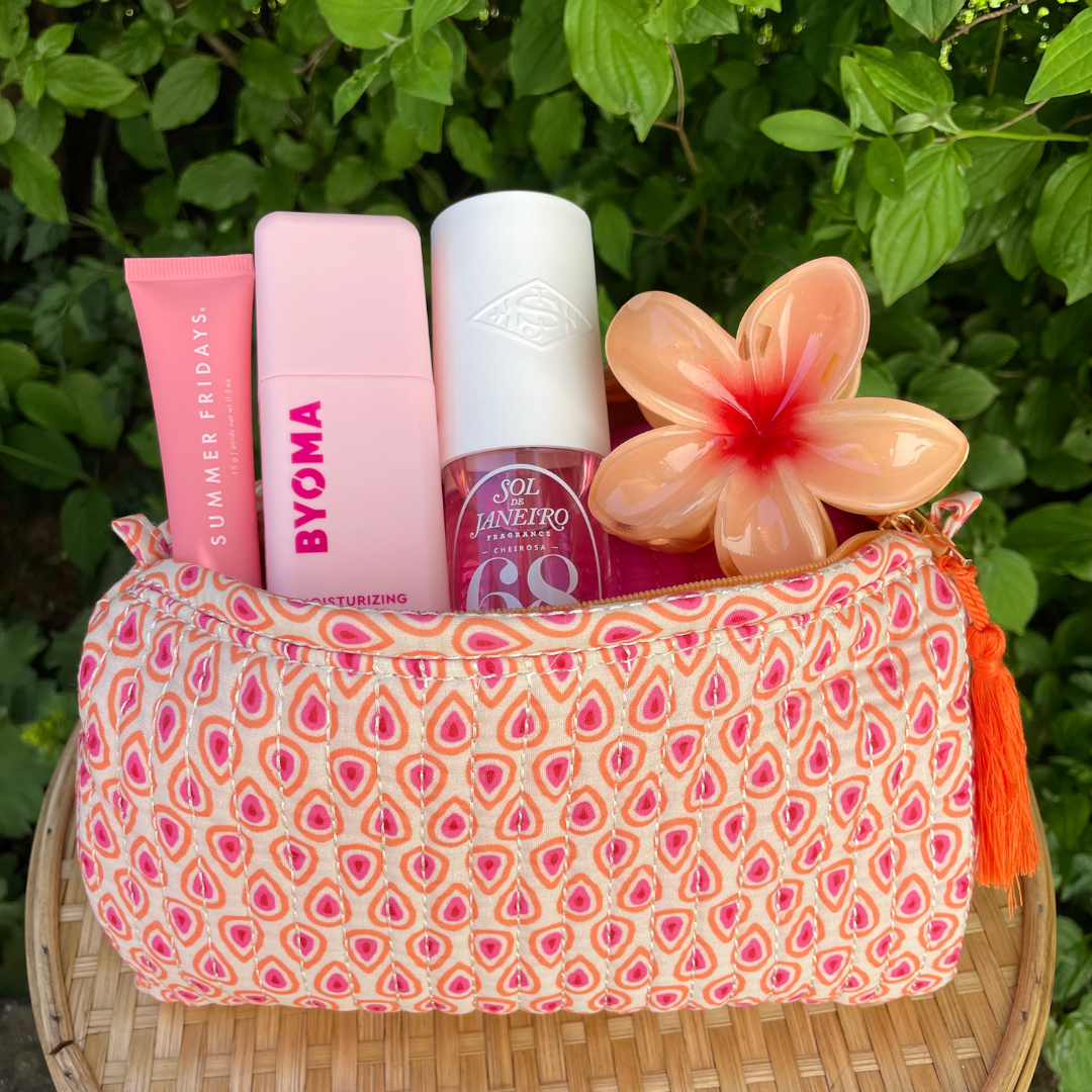 Gepolsterte Kosmetiktasche mit orange-rosa Tropfenmuster und orangefarbener Quaste, gefüllt mit Pflegeprodukten in Rosa sowie einer glänzenden pfirsichfarbenen Blumen-Haarklammer. Abgestellt auf einem runden Rattantisch vor grünem Pflanzenhintergrund. 
