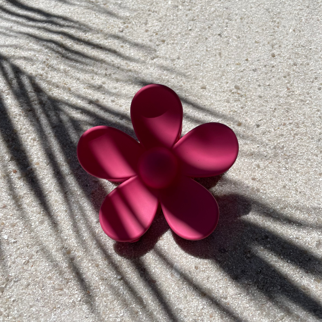 Haarklammer in Pink in Form einer fünfblättrigen Blume, arrangiert auf hellem Sand mit Schatten von Palmblättern im Hintergrund. Das sommerliche Accessoire setzt farbenfrohe Akzente im Haar. 