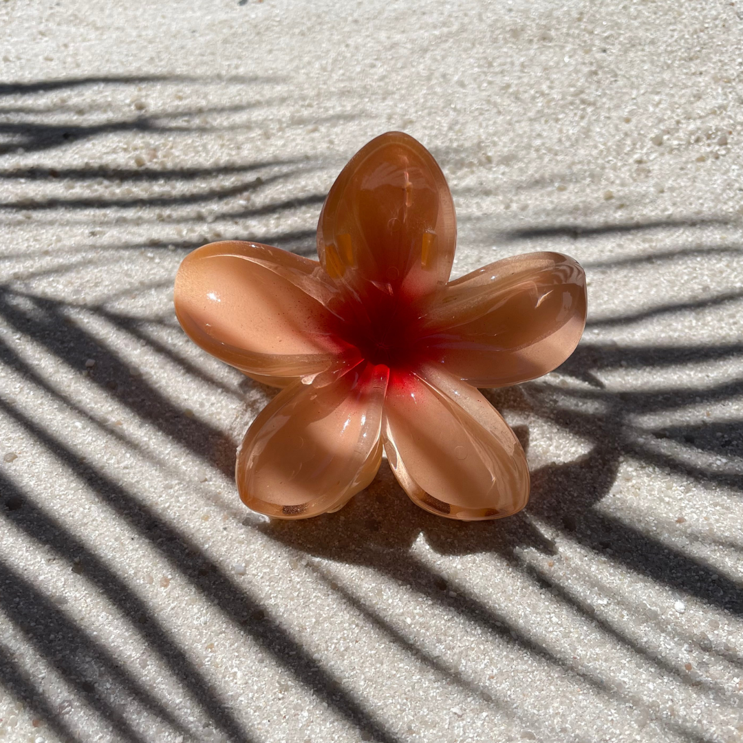 Blumenförmige Haarklammer in Apricot mit rotem Farbverlauf in der Mitte, arrangiert auf sandigem Untergrund im Sonnenlicht. Die Klammer erinnert an eine tropische Blüte und sorgt für einen sommerlichen Akzent im Haar. 