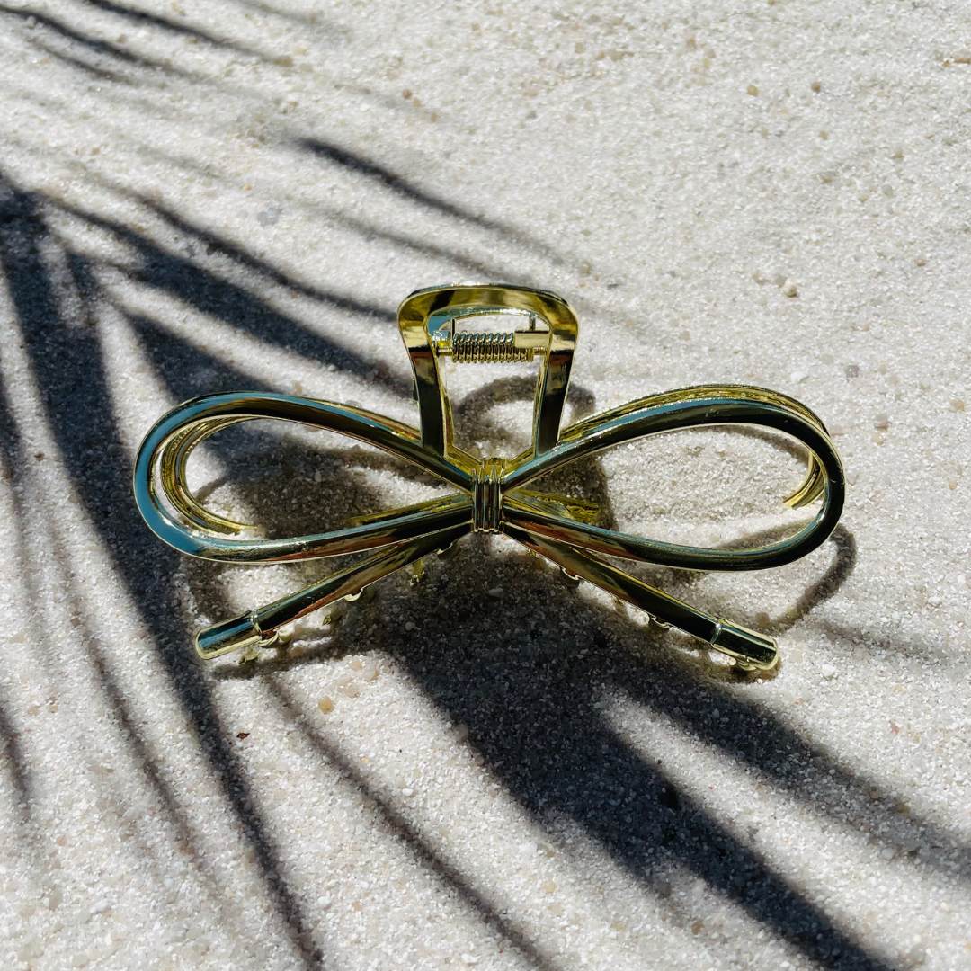Goldene Haarklammer aus Metall in Form einer Schleife, dekorativ auf hellem Sand platziert, umgeben von Palmenschatten. 