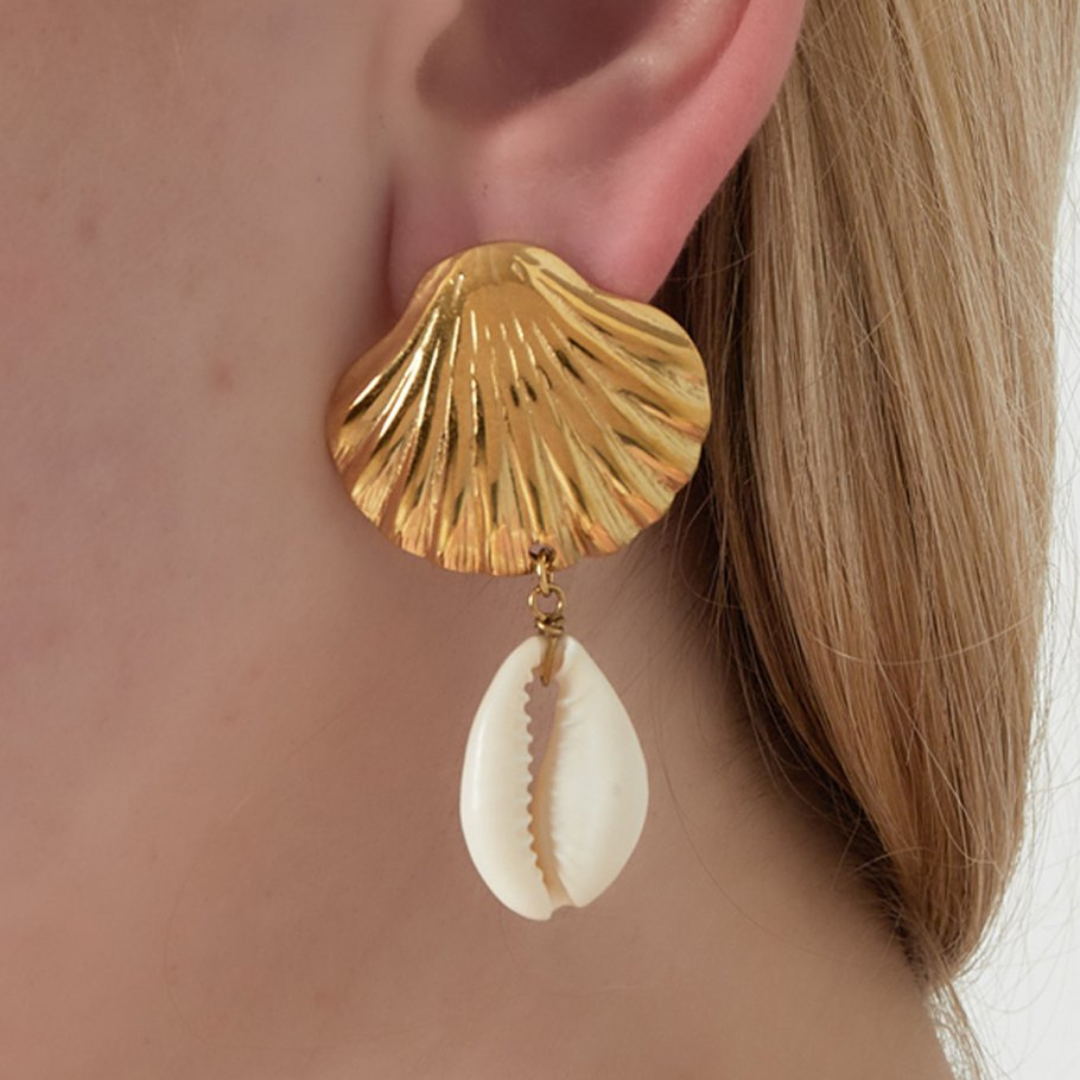 Goldfarbene Ohrringe in Muschelform mit echten Kaurimuscheln, getragen am Ohr vor hellem, blonden Haar. 