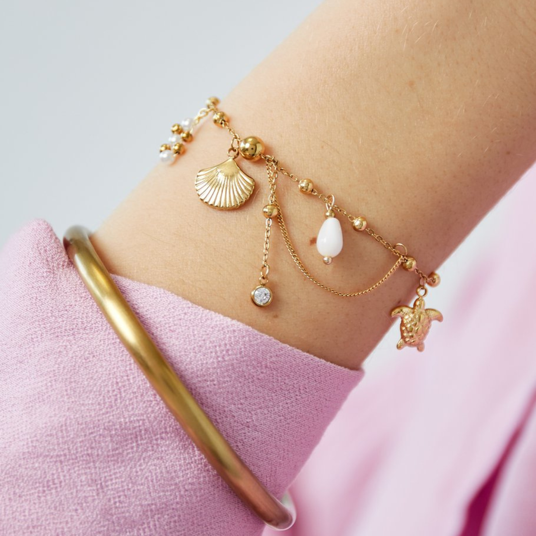 Goldfarbenes Armband mit verschiedenen Anhängern, z.B. Muschelanhänger, Perle, Zirkonia-Stein und feinen Kettendetails - getragen am Handgelenk, fotografiert im Studio mit Studiolicht. 