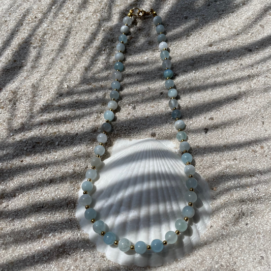 Halskette aus runden Aquamarin-Perlen mit kleinen goldfarbenen Kugeln dazwischen. Dekorativ auf einer weißen Muschelschale im Sand platziert. Im Sonnenlicht mit Palmenschatten. 