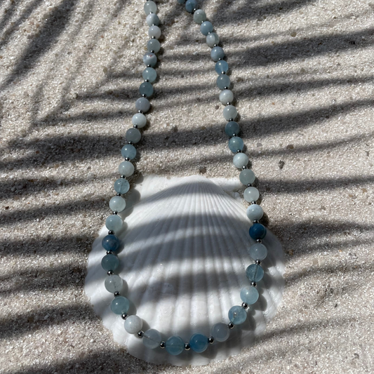 Halskette aus runden Aquamarin-Perlen mit kleinen silberfarbenen Kugeln dazwischen. Dekorativ auf einer weißen Muschelschale im Sand platziert. Im Sonnenlicht mit Palmenschatten. 