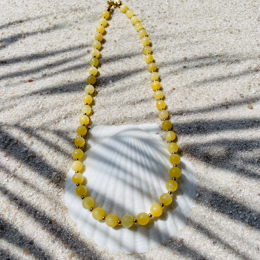 Halskette aus runden gelben Jadeperlen mit kleinen goldenen Kugeln dazwischen. Dekorativ auf einer weißen Muschelschale im Sand platziert. Im Sonnenlicht mit Palmenschatten. 