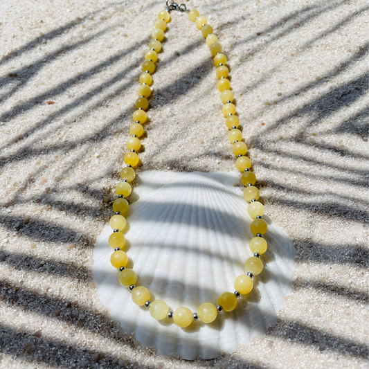 Halskette aus runden gelben Jadeperlen mit kleinen silberfarbenen Kugeln dazwischen. Dekorativ auf einer weißen Muschelschale im Sand platziert. Im Sonnenlicht mit Palmenschatten. 