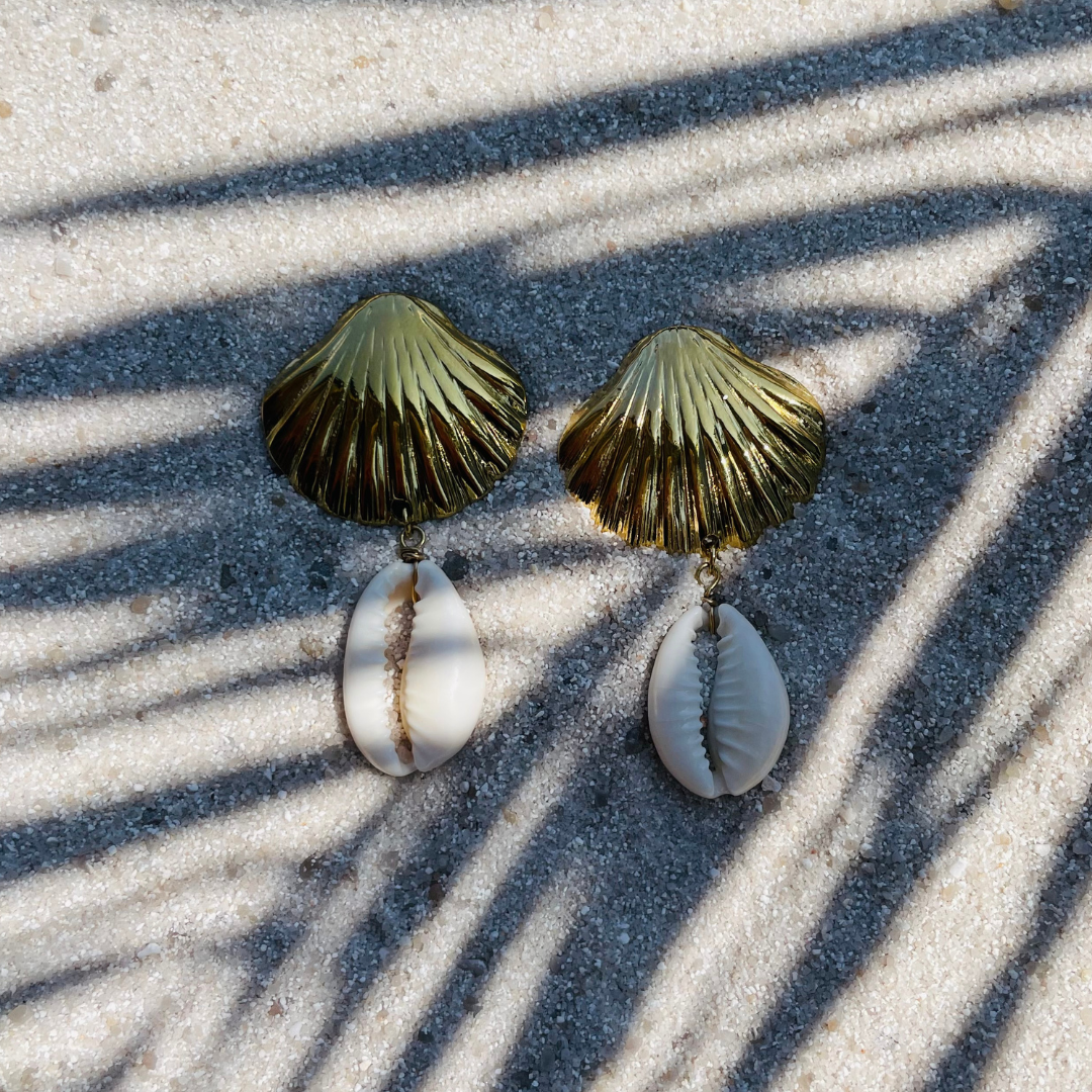 Goldfarbene Ohrringe in Muschelform mit echten Kaurimuscheln, arrangiert auf Sand mit Palmenschatten im Hintergrund. 