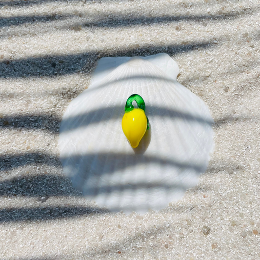 Ein Zitronen Charm aus Glas, mit leuchtend gelber Frucht und grünem Blattansatz. Der Anhänger liegt dekorativ auf einer weißen Muschelschale im Sand. Sonnenlicht und Palmenschatten erzeugen ein sommerliches, maritimes Ambiente. 