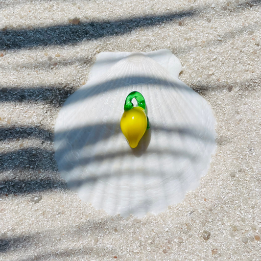 Ein Zitronen Charm aus Glas, mit leuchtend gelber Frucht und grünem Blattansatz. Der Anhänger liegt dekorativ auf einer weißen Muschelschale im Sand. Sonnenlicht und Palmenschatten erzeugen ein sommerliches, maritimes Ambiente. 