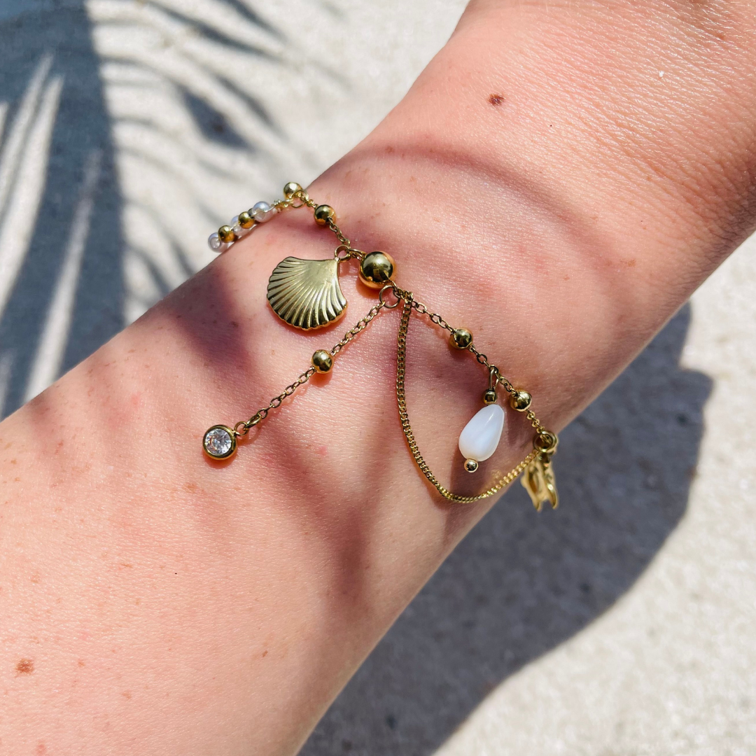 Goldfarbenes Armband mit verschiedenen Anhängern, z.B. Muschelanhänger, Perle, Zirkonia-Stein und  Kettendetails - getragen am Handgelenk, fotografiert bei Sonnenlicht auf sandigem Untergrund. 