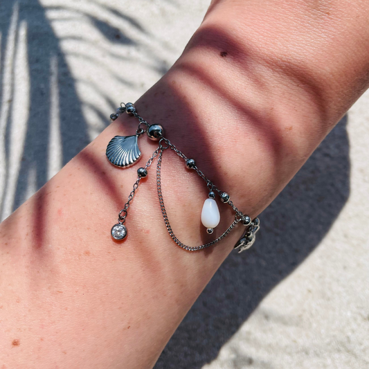 Silberfarbenes Armband mit verschiedenen Anhängern, z.B. Muschelanhänger, Perle, Zirkonia-Stein und feinen Kettendetails - getragen am Handgelenk, fotografiert bei Sonnenlicht auf sandigem Untergrund. 
