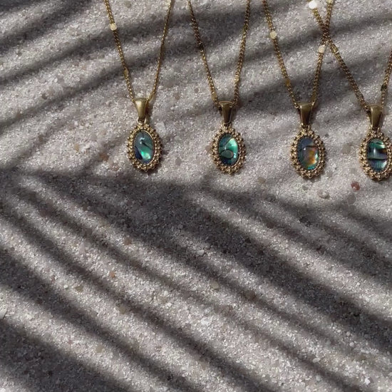 Im Video sieht man fünf goldene Halsketten mit ovalen Anhängern aus Abalone-Perlmutt, die nebeneinander auf hellem Sand liegen. Durch das Licht erkennt man deutlich die schimmernden Muster - jede Kette ist ein Unikat. 