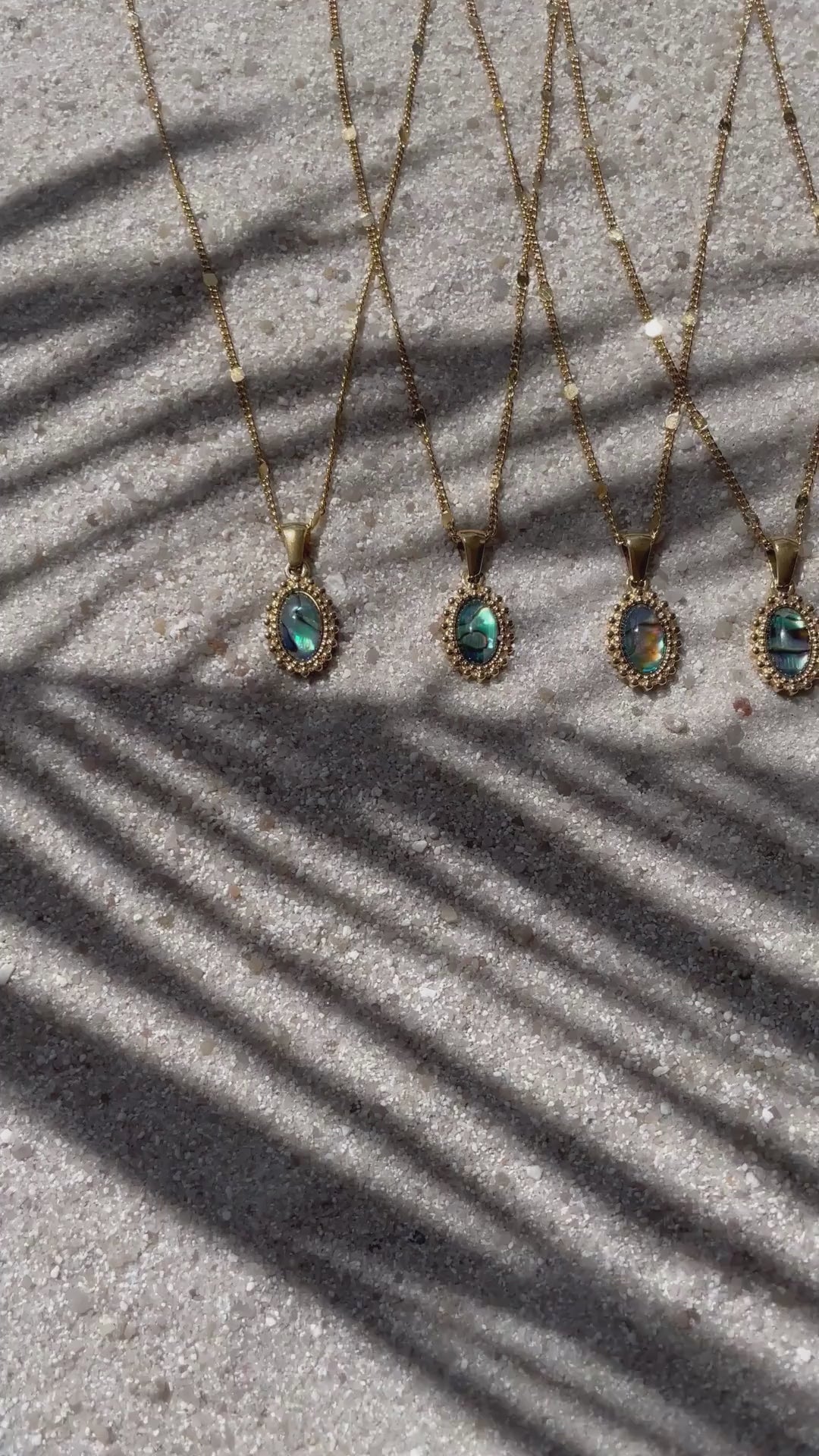 Im Video sieht man fünf goldene Halsketten mit ovalen Anhängern aus Abalone-Perlmutt, die nebeneinander auf hellem Sand liegen. Durch das Licht erkennt man deutlich die schimmernden Muster - jede Kette ist ein Unikat. 
