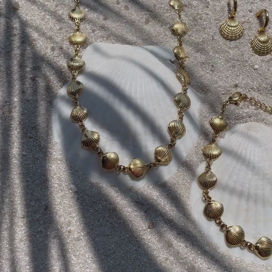 Video mit Goldfarbenem Schmuckset bestehend aus Halskette, Armband und Creolen - alle mit kleinen Jakobsmuschel-Anhängern. Das Set liegt auf Sand, Sonnenlicht und Schatten bewegen sich sanft über die Schmuckstücke. 