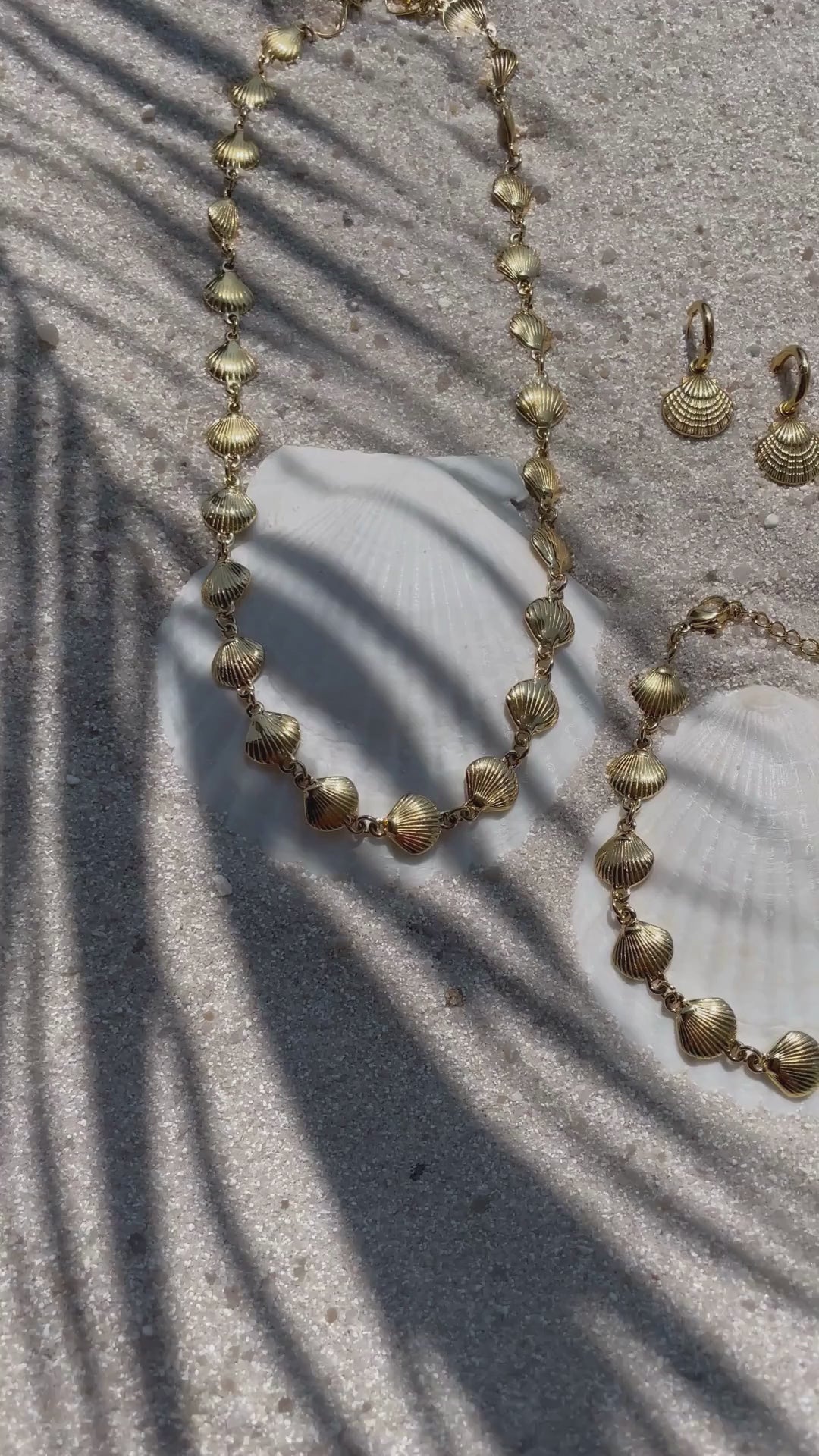 Video mit Goldfarbenem Schmuckset bestehend aus Halskette, Armband und Creolen - alle mit kleinen Jakobsmuschel-Anhängern. Das Set liegt auf Sand, Sonnenlicht und Schatten bewegen sich sanft über die Schmuckstücke. 