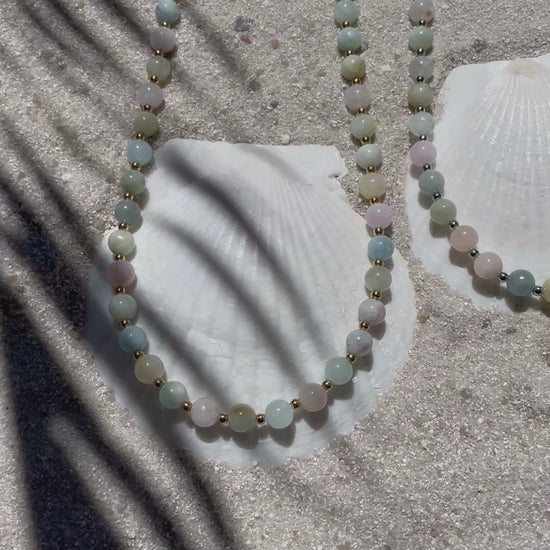 Zwei handgefertigte Halsketten aus pastellfarbenen Beryll-Perlen - eine mit kleinen goldfarbenen, die andere mit kleinen silberfarbenen Kugeln dazwischen. Präsentiert auf feinem Sand mit weißen Muscheln, im natürlichen Sonnenlicht mit zarten Schattenspielen.