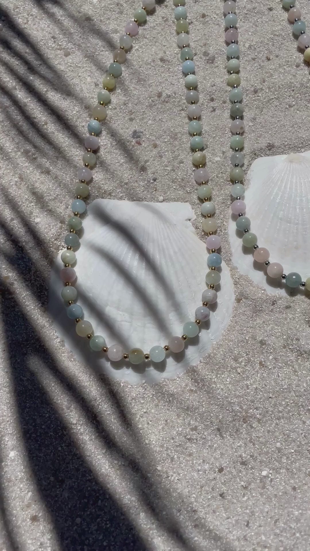 Zwei handgefertigte Halsketten aus pastellfarbenen Beryll-Perlen - eine mit kleinen goldfarbenen, die andere mit kleinen silberfarbenen Kugeln dazwischen. Präsentiert auf feinem Sand mit weißen Muscheln, im natürlichen Sonnenlicht mit zarten Schattenspielen.