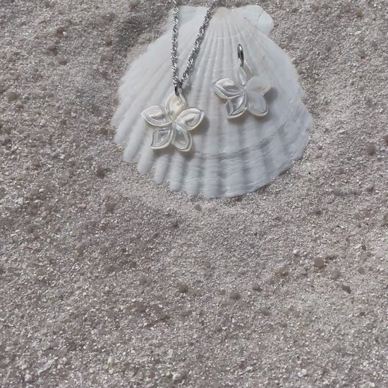 Ein schimmernder Blütenanhänger aus weißer Muschelperle liegt neben einer passenden Halskette auf einer weißen Muschelschale im Sand. Die filigrane Kette ist silberfarben und spiegelt das Licht. 