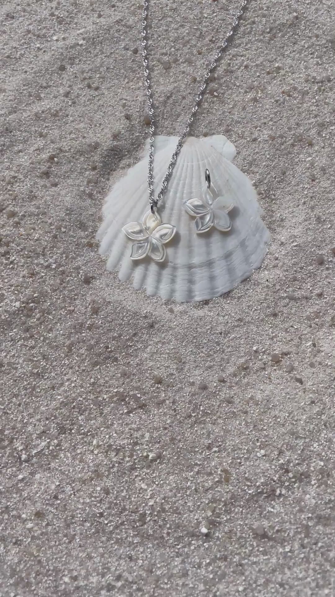 Ein schimmernder Blütenanhänger aus weißer Muschelperle liegt neben einer passenden Halskette auf einer weißen Muschelschale im Sand. Die filigrane Kette ist silberfarben und spiegelt das Licht. 