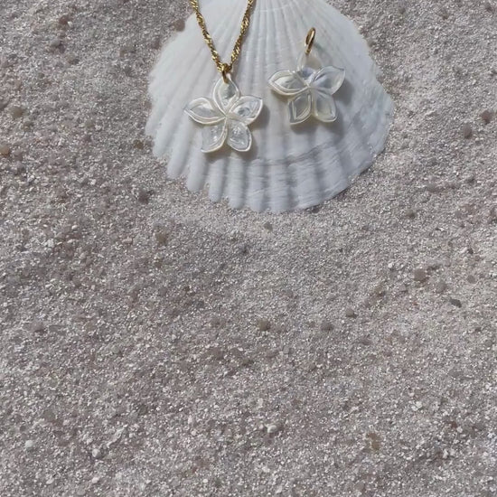 Ein schimmernder Blütenanhänger aus weißer Muschelperle liegt neben einer passenden Halskette auf einer weißen Muschelschale im Sand. Die filigrane Kette ist goldfarben und ist aus wasserfestem und vergoldeten Edelstahl. 