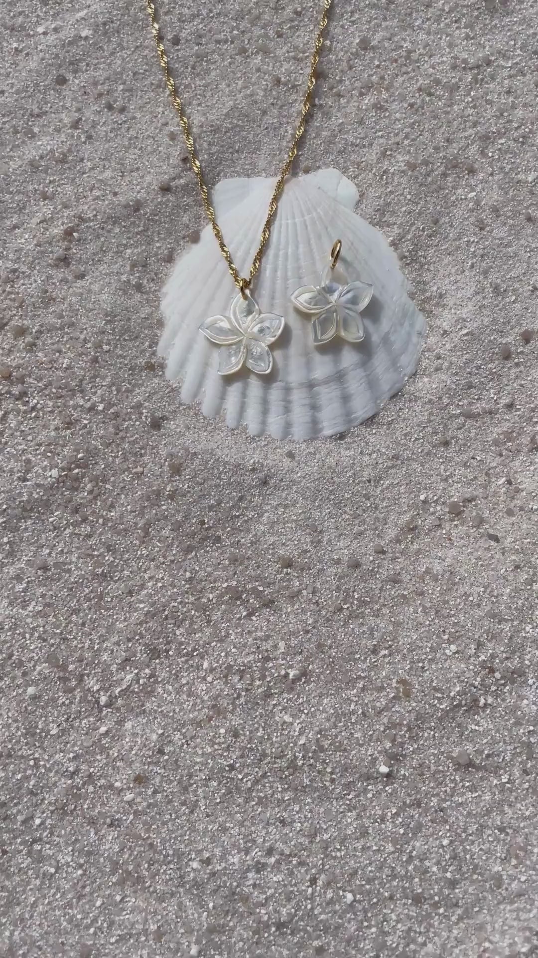 Ein schimmernder Blütenanhänger aus weißer Muschelperle liegt neben einer passenden Halskette auf einer weißen Muschelschale im Sand. Die filigrane Kette ist goldfarben und ist aus wasserfestem und vergoldeten Edelstahl. 